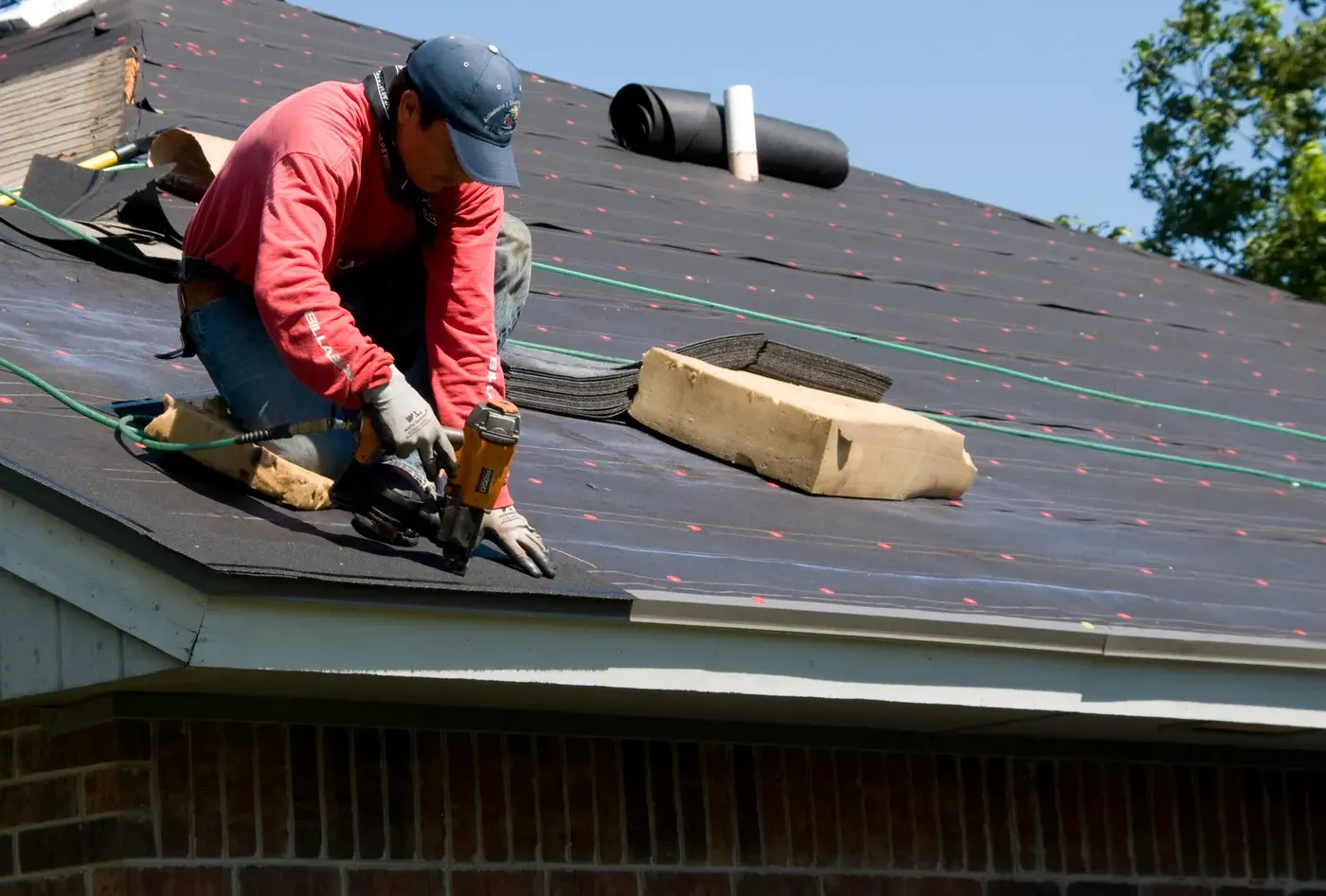 A roofer kneeling at the eave, fastening a new course of shingles above the gutter line